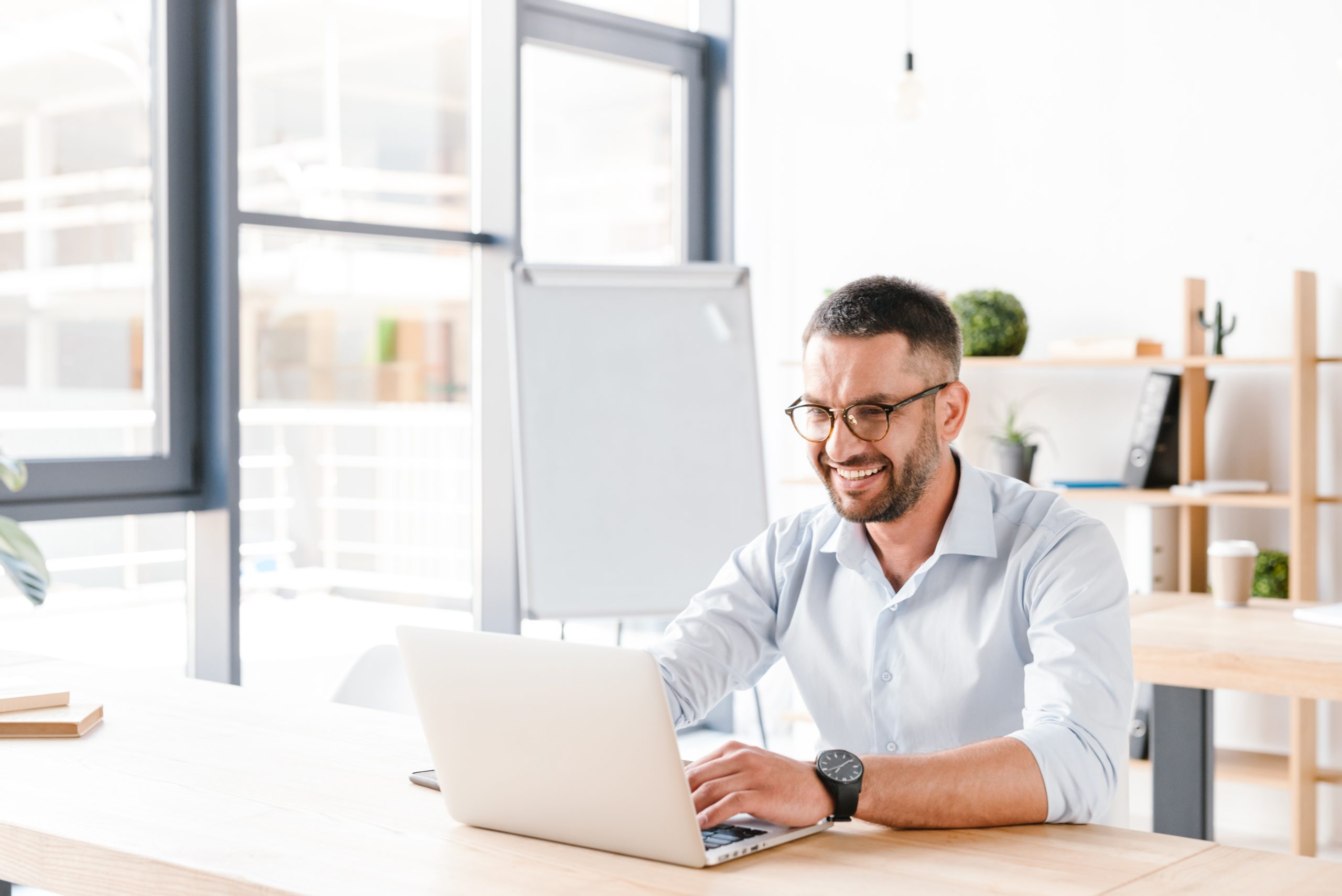 Foto eines fröhlichen Büromenschen in den 30ern in weißem Hemd, der am Schreibtisch sitzt und an einem Laptop in einem Geschäftszentrum arbeitet