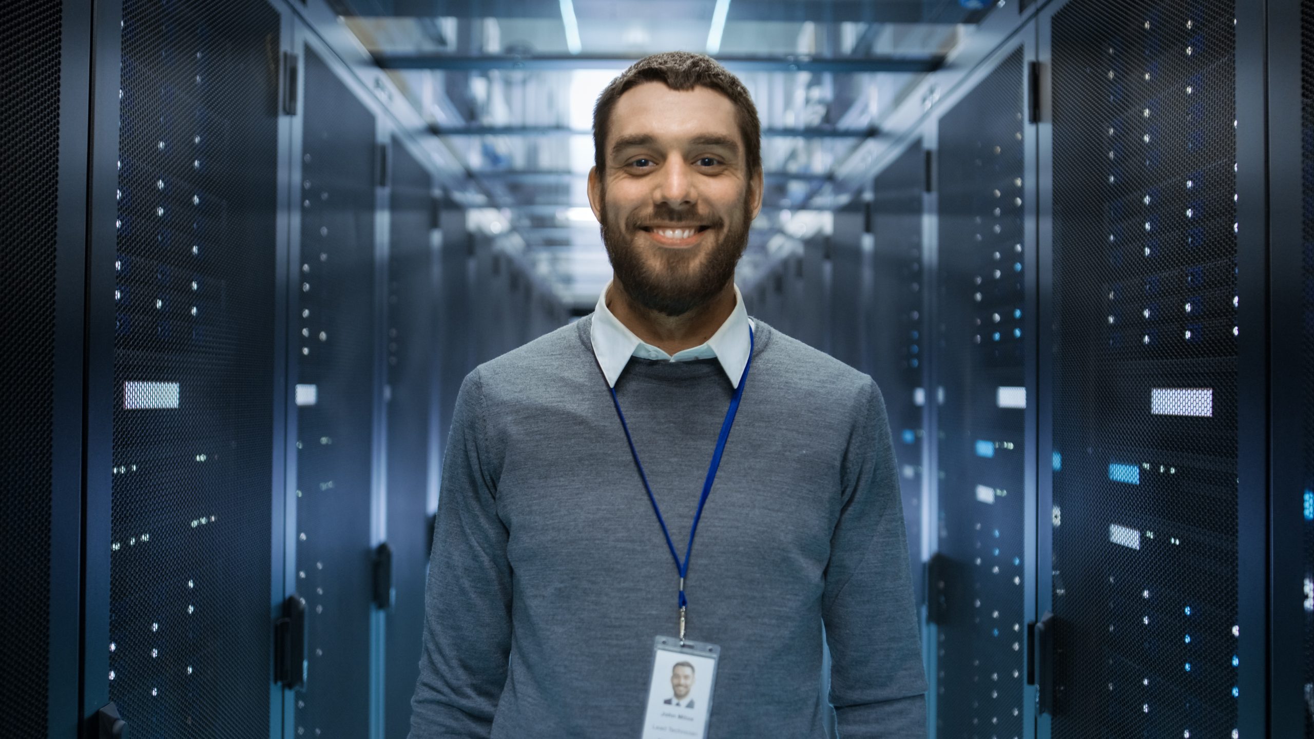 Porträt eines neugierigen, positiv gestimmten und lächelnden IT-Ingenieurs, der in der Mitte eines großen Serverraums im Rechenzentrum steht.